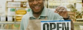 smiling man holding a sign that the business is open thanks to small business grants